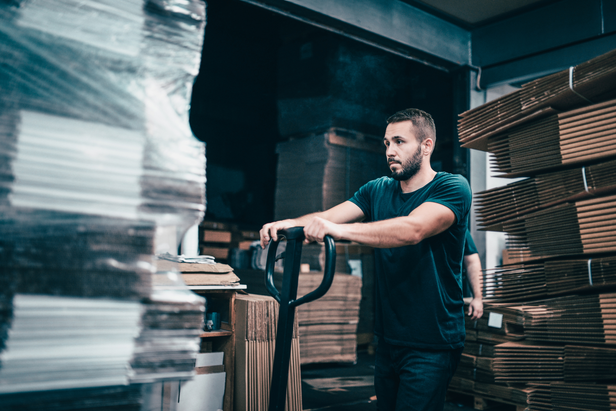 Worker In Cardboard Factory Pushing Pallets On To Transportation Cart