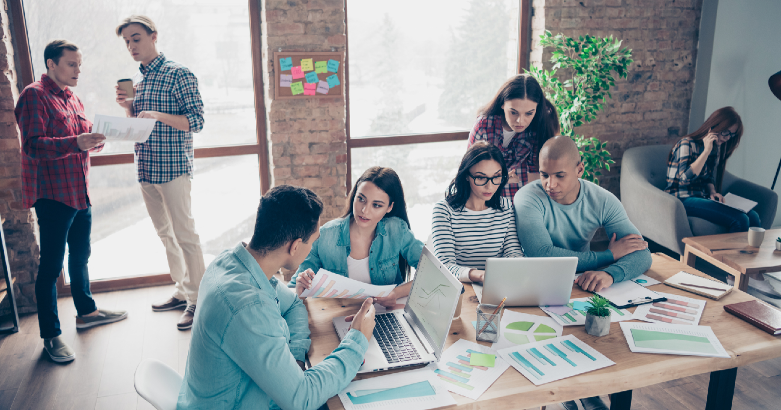 employees gathered in a workspace at a table sharing reports, looking at laptops, and talking.